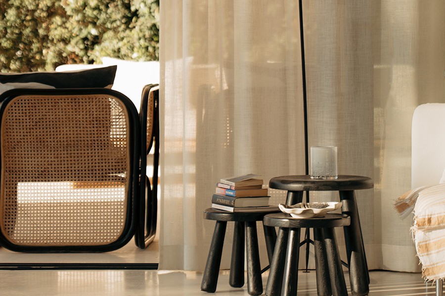 Rattan armchair beside sheer curtains, with nested stools styled with books and a candle